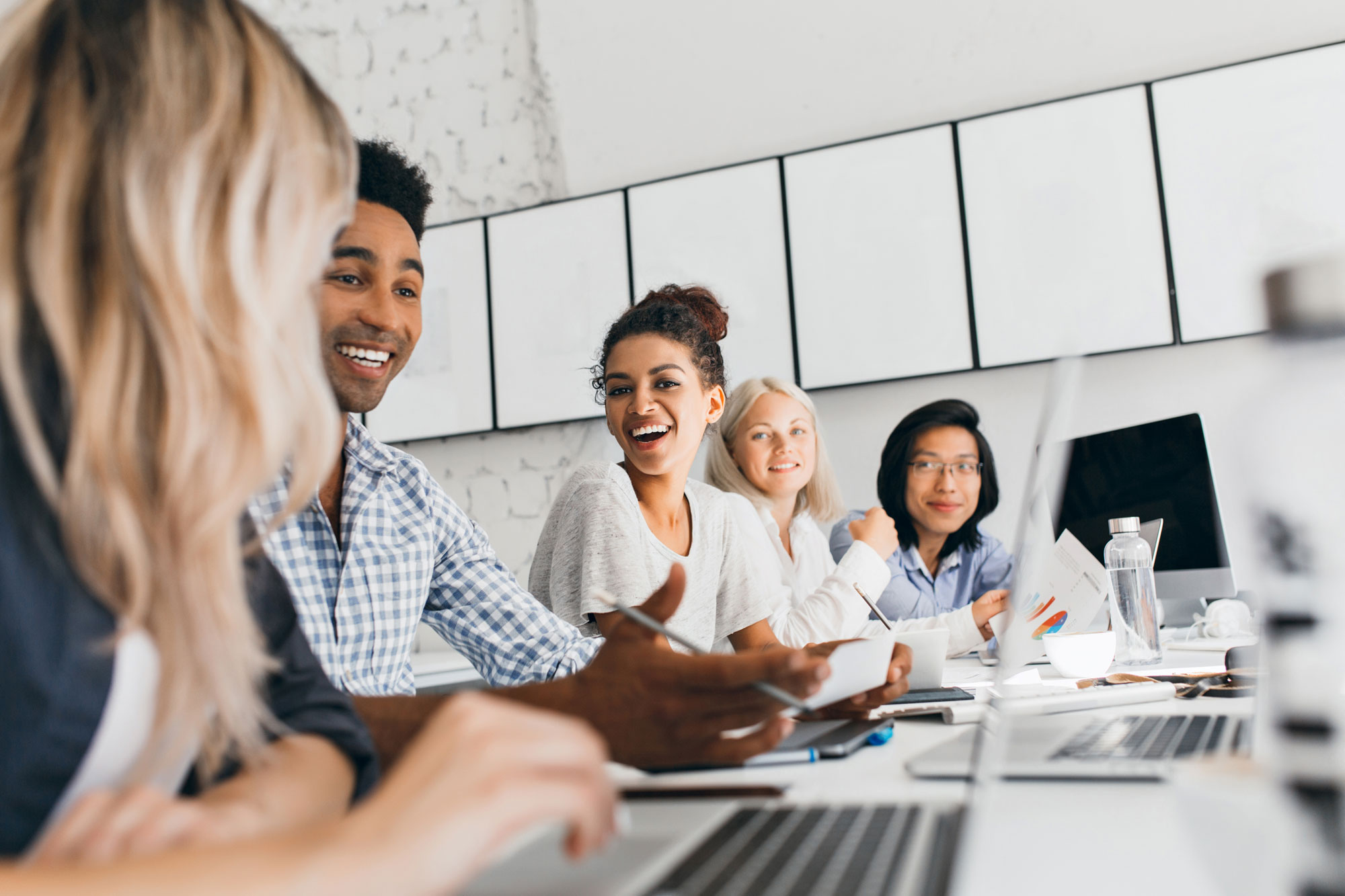 A diverse group of people sit around a table with laptops, papers, and a water bottle, smiling as they engage in conversation. In the bright room, where non-proprietary access control hardware ensures seamless collaboration, a series of blank canvases adorns the wall in the background.