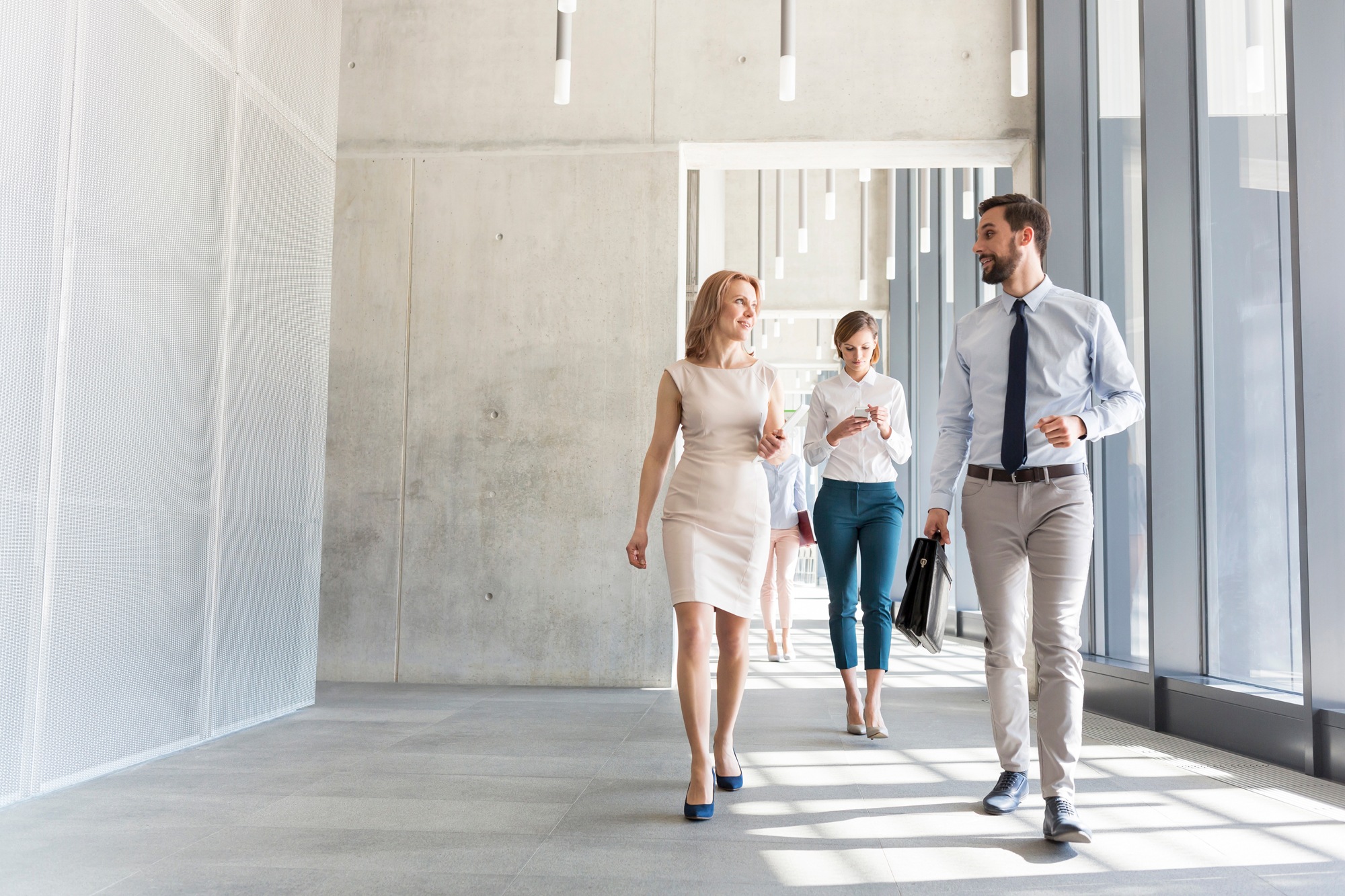 Three business professionals are walking in a modern office hallway, discussing process automation. A woman in a beige dress and a man in a light shirt and tie lead the conversation. Another woman in a white shirt and teal pants follows, holding a phone. Large windows line one side.