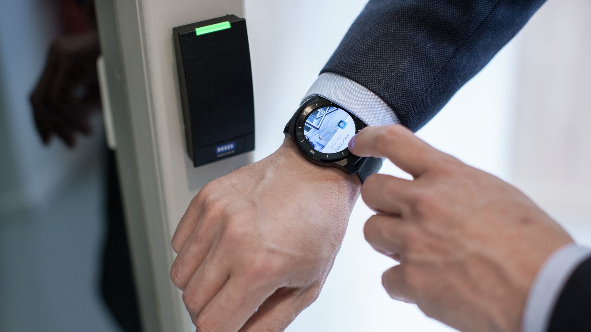 A person wearing a smartwatch holds their wrist near a security card reader on a door, engaging in access control lingo. Dressed in formal attire, the individual stands in an indoor setting, suggesting verification of identity.