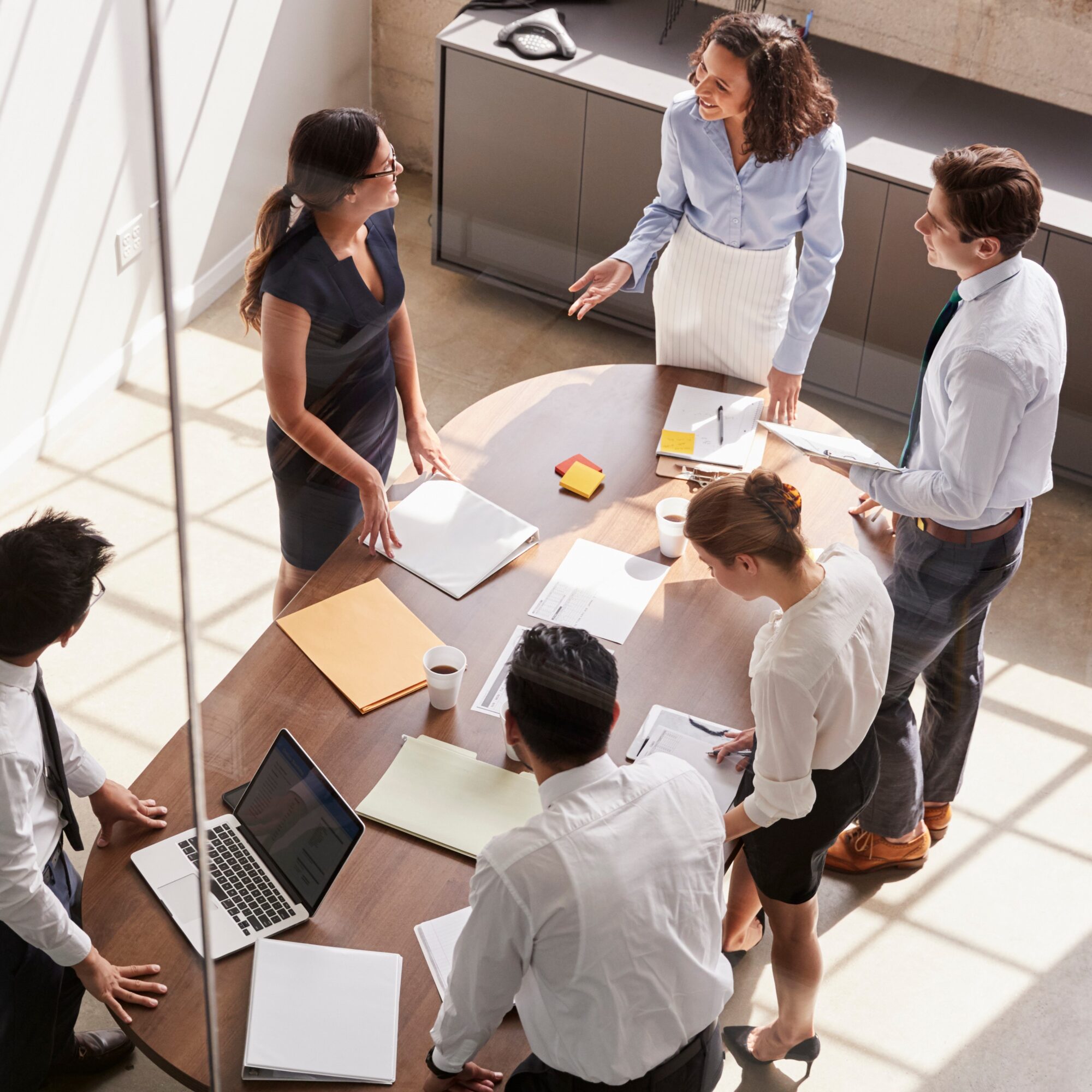 A diverse group of professionals stands around a table in an office, engaged in a collaborative discussion about the latest BMS-agnostic On-Demand HVAC solution. Laptops, notebooks, and coffee cups are on the table. A woman in a blue shirt gestures as she speaks, while others listen attentively.