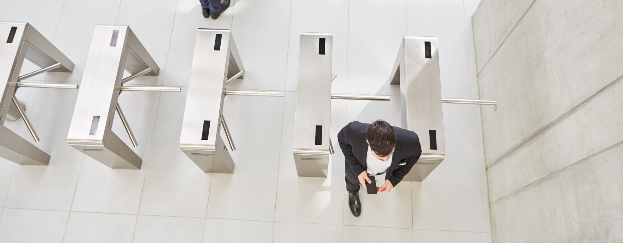 A person in formal attire walks through a modern turnstile in a spacious, bright area equipped with advanced access control system software. The floor is tiled, and there is another visible turnstile nearby. A small section of another person is seen in the background approaching a turnstile.