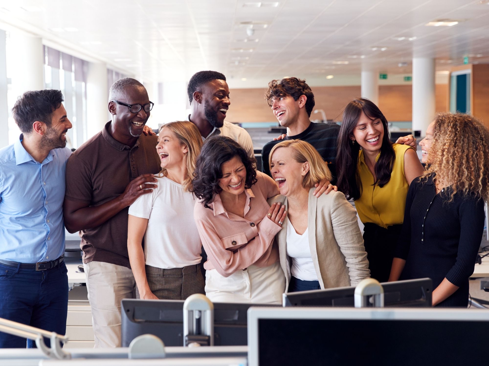 A diverse group of colleagues in casual business attire share a laugh in an open Genea Careers office space, with desks and computers visible in the foreground. They appear to be enjoying a light moment together.