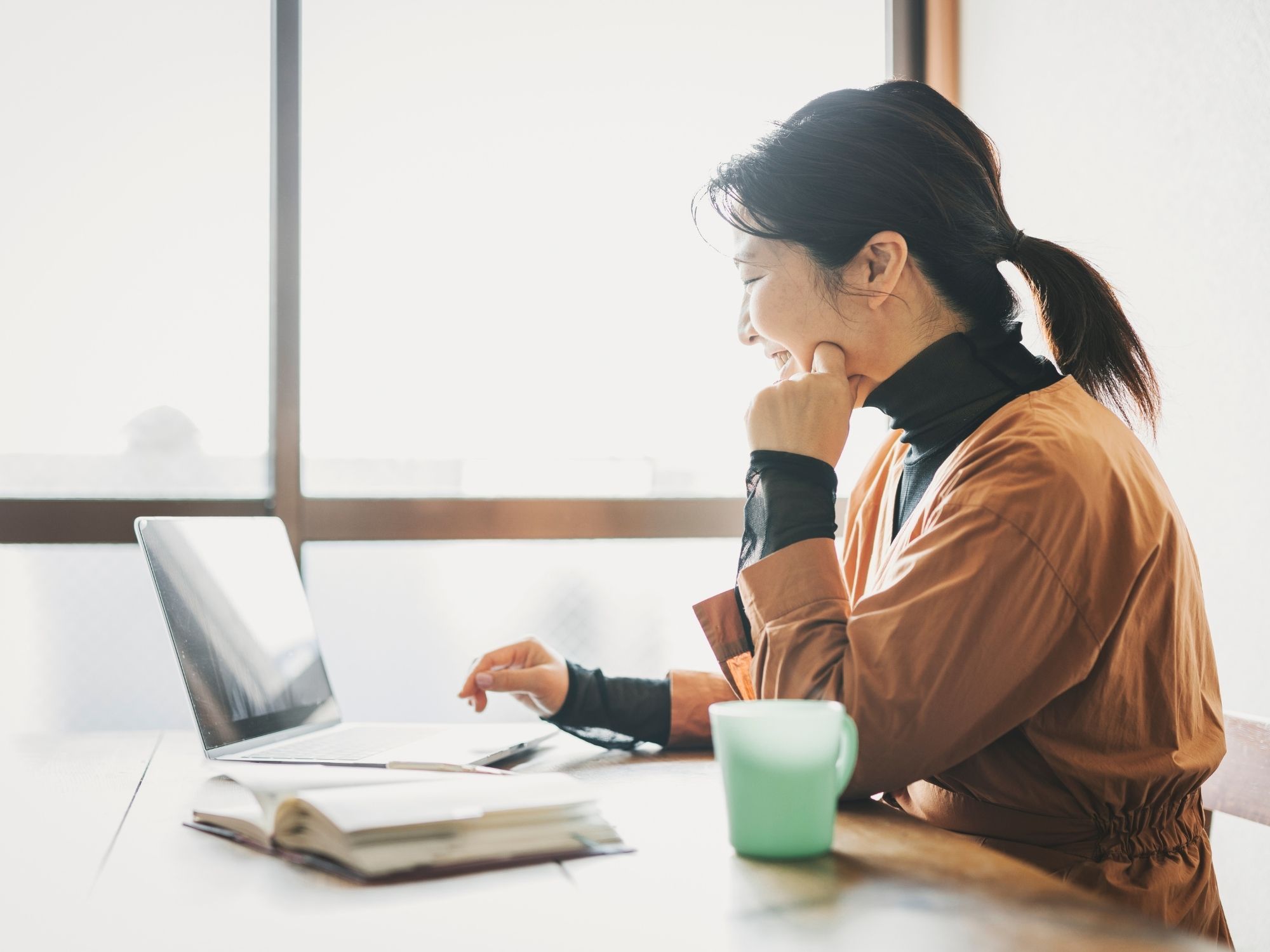 A woman in a brown jacket sits at a wooden table, smiling at her laptop screen as she explores exciting opportunities with Genea Careers. An open notebook and a green mug are in front of her, while sunlight streams through the window, creating a warm, inviting atmosphere.