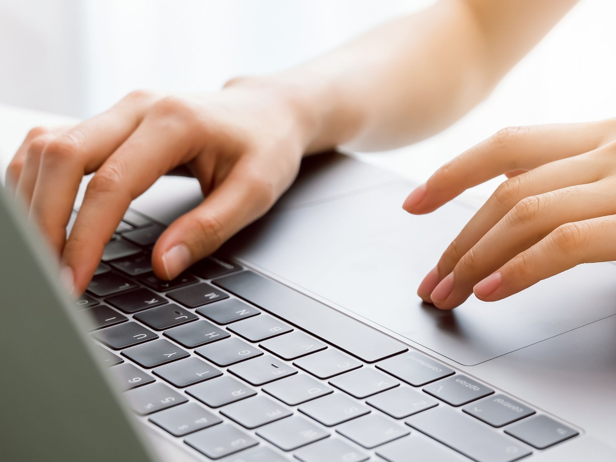 Close-up of a person typing on a sleek, modern laptop keyboard, suggesting they're immersed in exploring Genea careers. The hands press the keys with purpose against a bright, blurred background, indicating a well-lit indoor setting conducive to focused job searching.