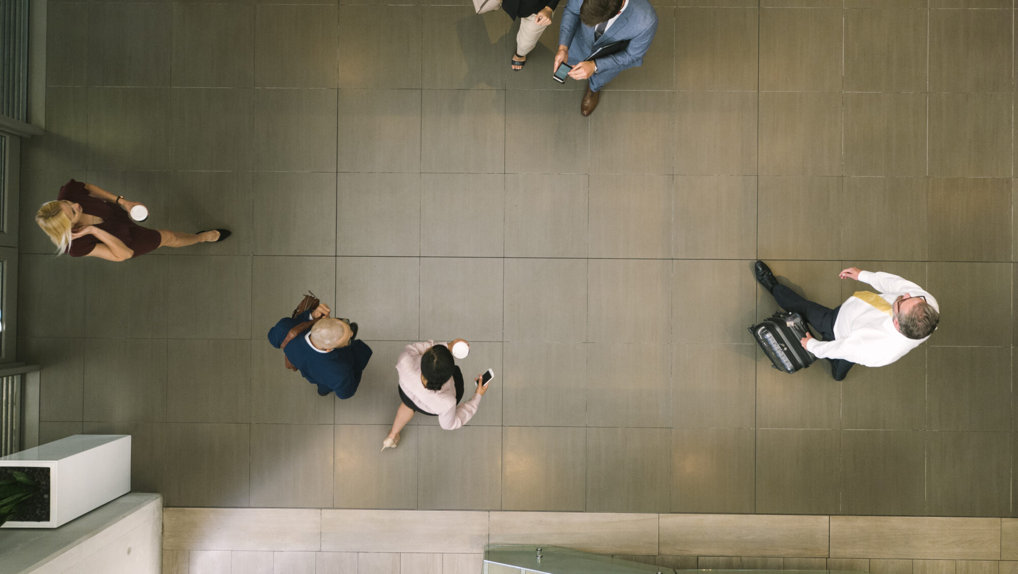 Aerial view of people walking on a tiled floor. Six individuals are scattered, some holding coffee cups or mobile devices with mobile credentials. One person pulls a suitcase. They are dressed in business attire.
