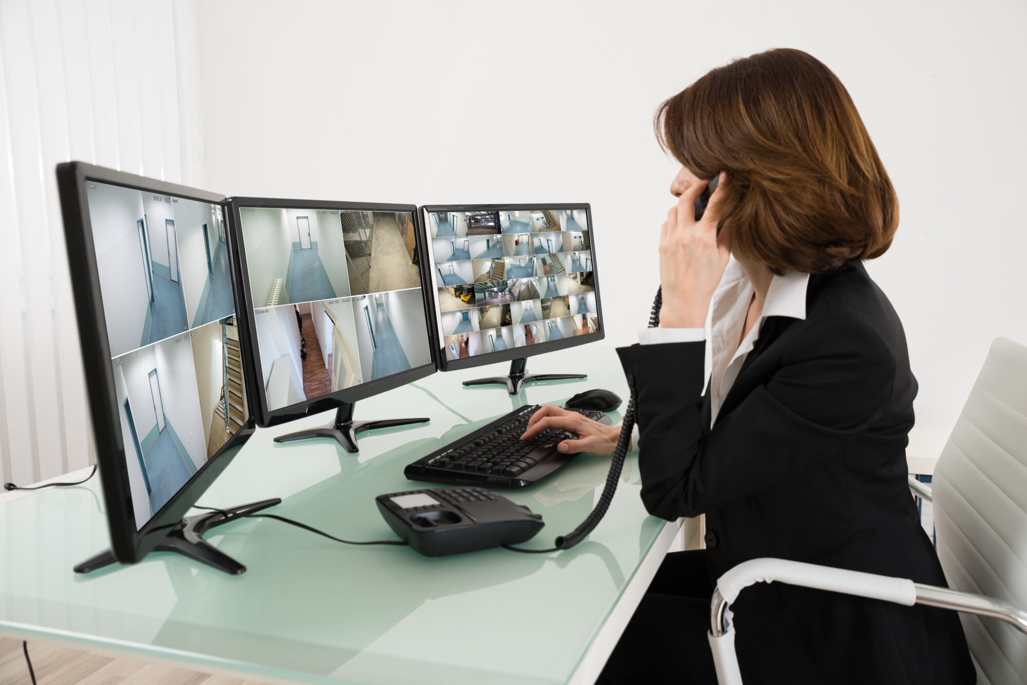 A woman in a black suit sits at a desk with three monitors displaying surveillance camera feeds, seamlessly managing video operations. She is holding a phone to her ear and looking at the screens attentively, embodying the efficiency of a security monitoring room.