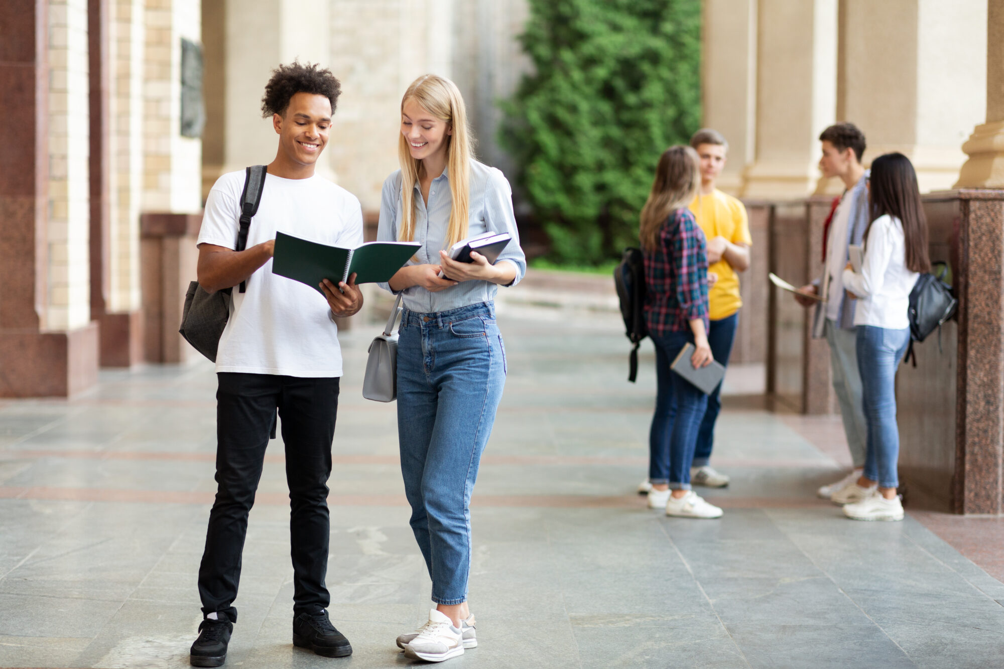 A group of students is standing in a college courtyard. In the foreground, a young man and woman are smiling while looking at a book. Others are chatting in the background, holding notebooks near the access control panel for schools. The scene is lively and studious.