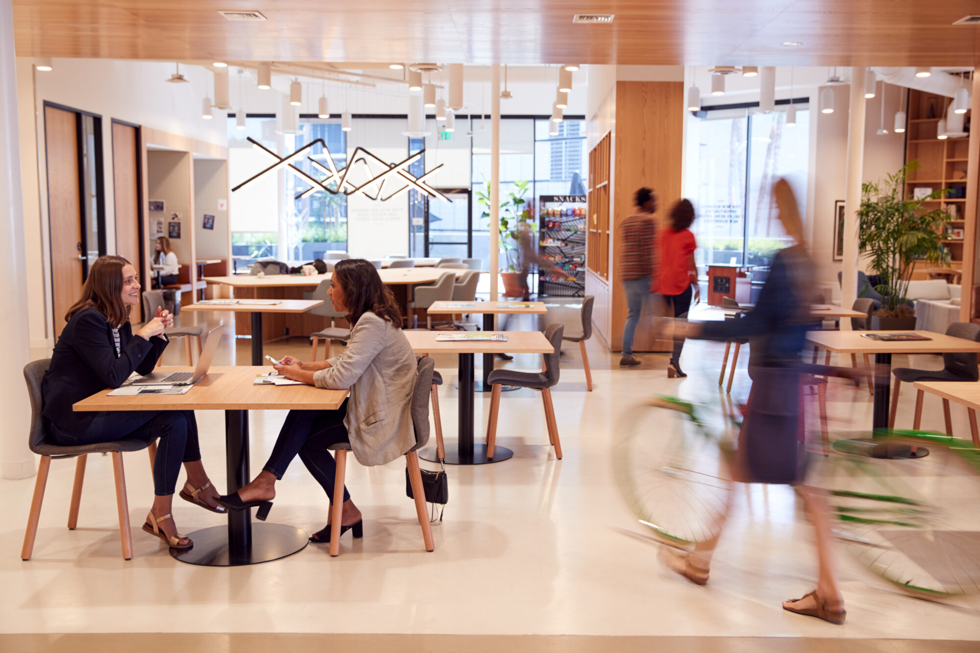 A modern coworking space with two people discussing work at a table, as others move about or work separately. A person rides a bicycle indoors, while the open space provides natural light and plants for decoration, ensuring both creativity and security are seamlessly integrated.