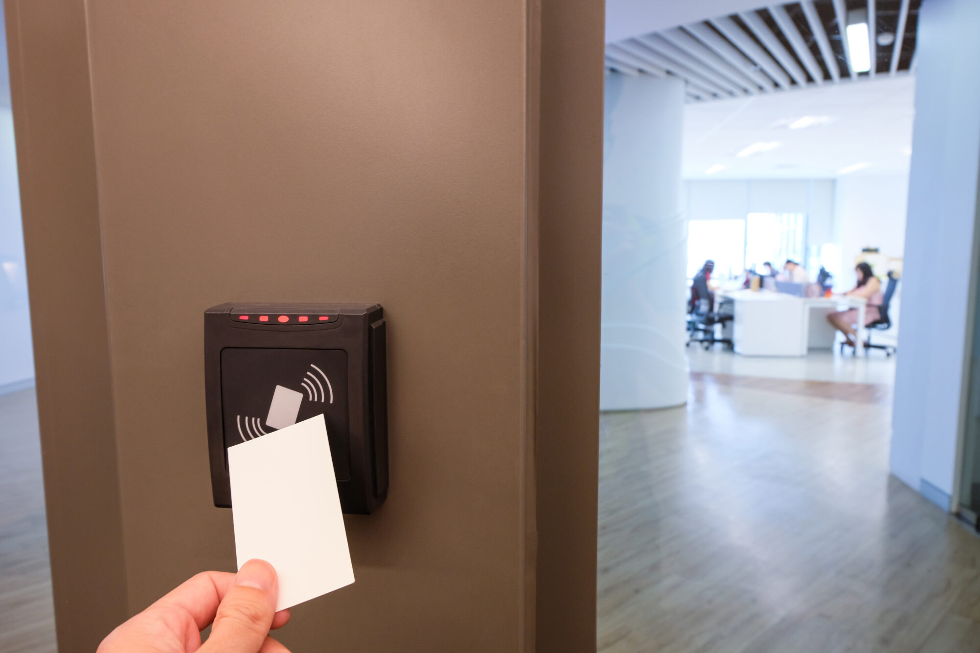 A hand holds a white access card in front of a card reader on the wall, showcasing an efficient small business access control system. In the background, an office environment is visible with several people seated at desks.