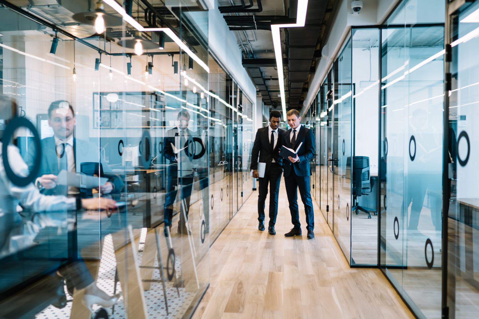 Two men in suits walk down a modern office corridor lined with glass-walled rooms, discussing and looking at a notebook. The hallway features sleek design elements like reflective surfaces and advanced door access control systems, while other people are visible through the glass.