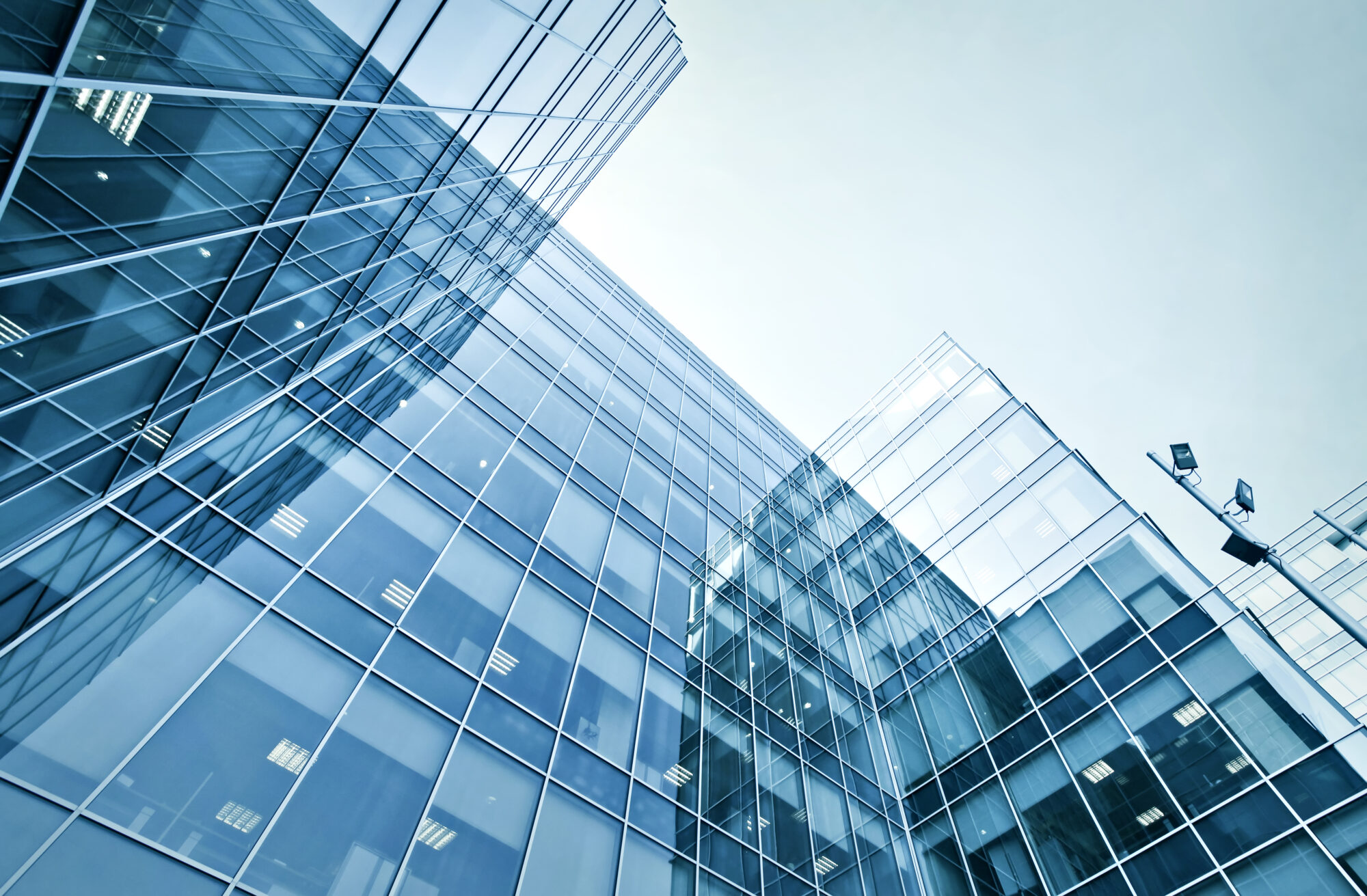 A low-angle view of a modern glass office building against a clear sky background. The building, home to an IT and Security firm, features reflective blue-tinted windows, capturing the surrounding urban environment and cloudless sky.
