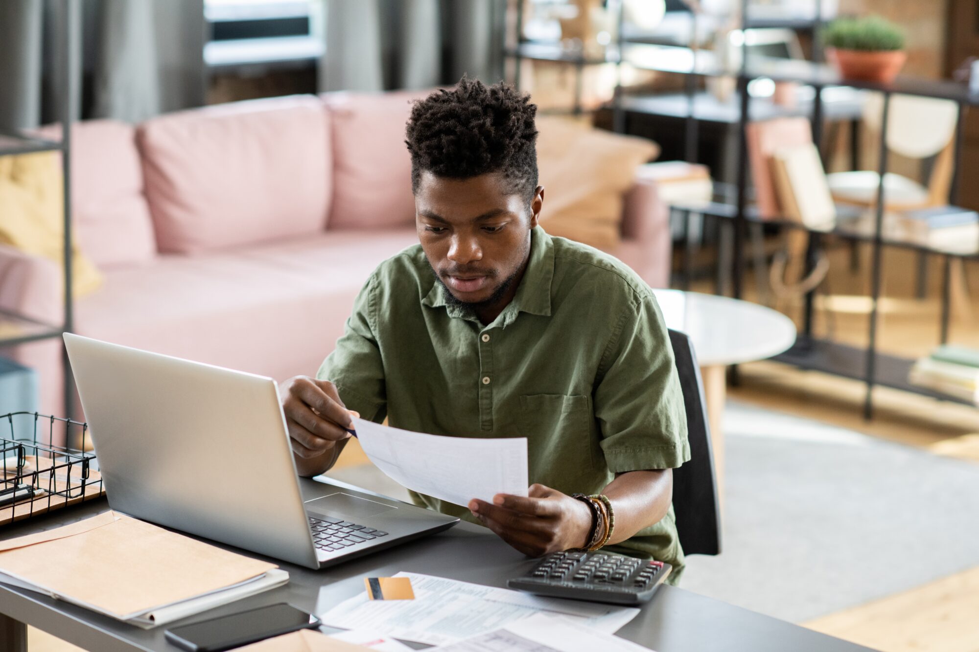 A building engineer sits at a desk, poring over a document in hand. A laptop is open and a calculator rests nearby. The cozy room features a sofa and bookshelves, adding to the studious atmosphere as they plan their next project.