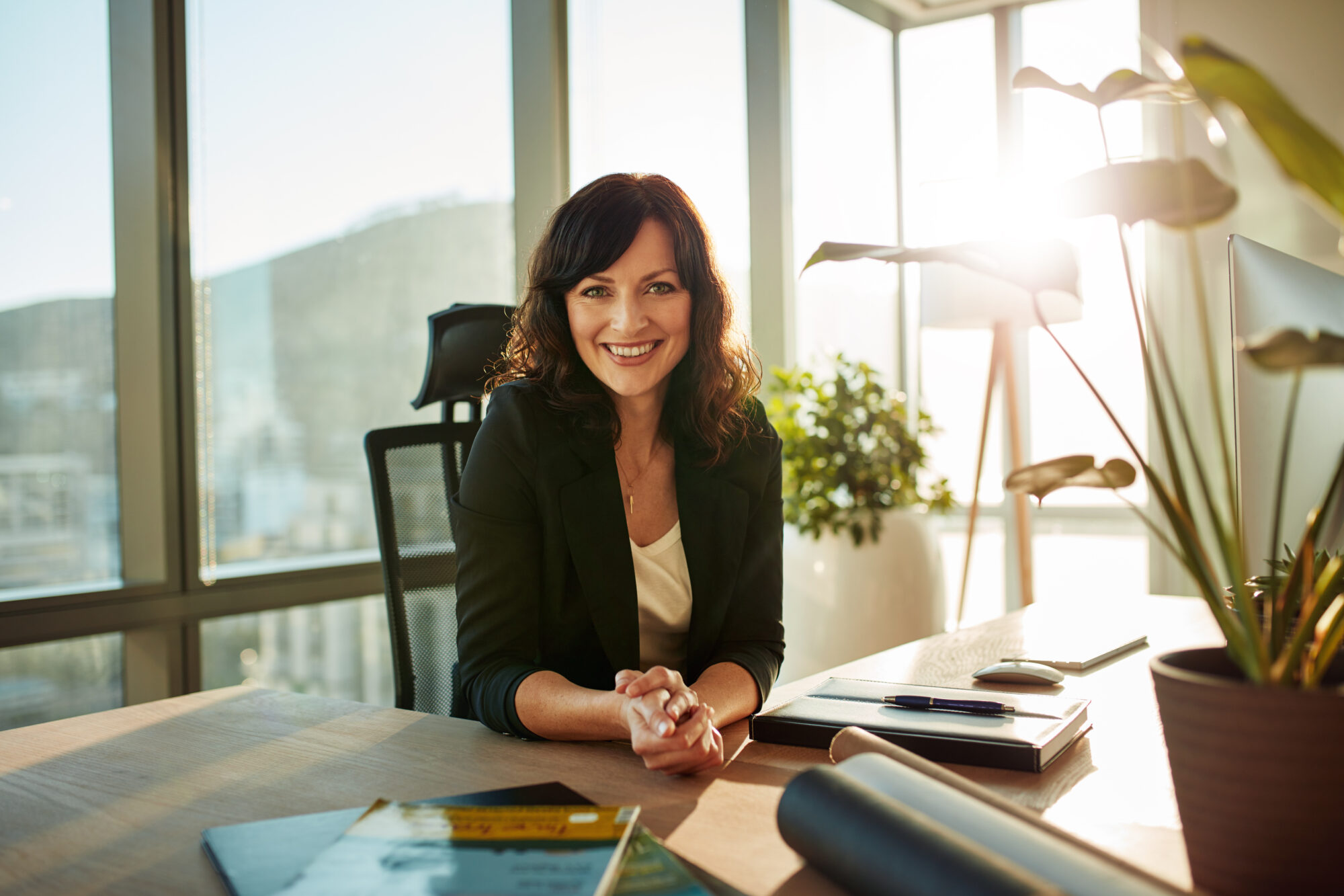 A woman with dark hair smiles at a desk in a sunlit office, embodying the confident aura of successful property managers. She wears a black blazer over a white shirt, while nearby are a plant and some documents. Large windows reveal an impressive cityscape in the background.