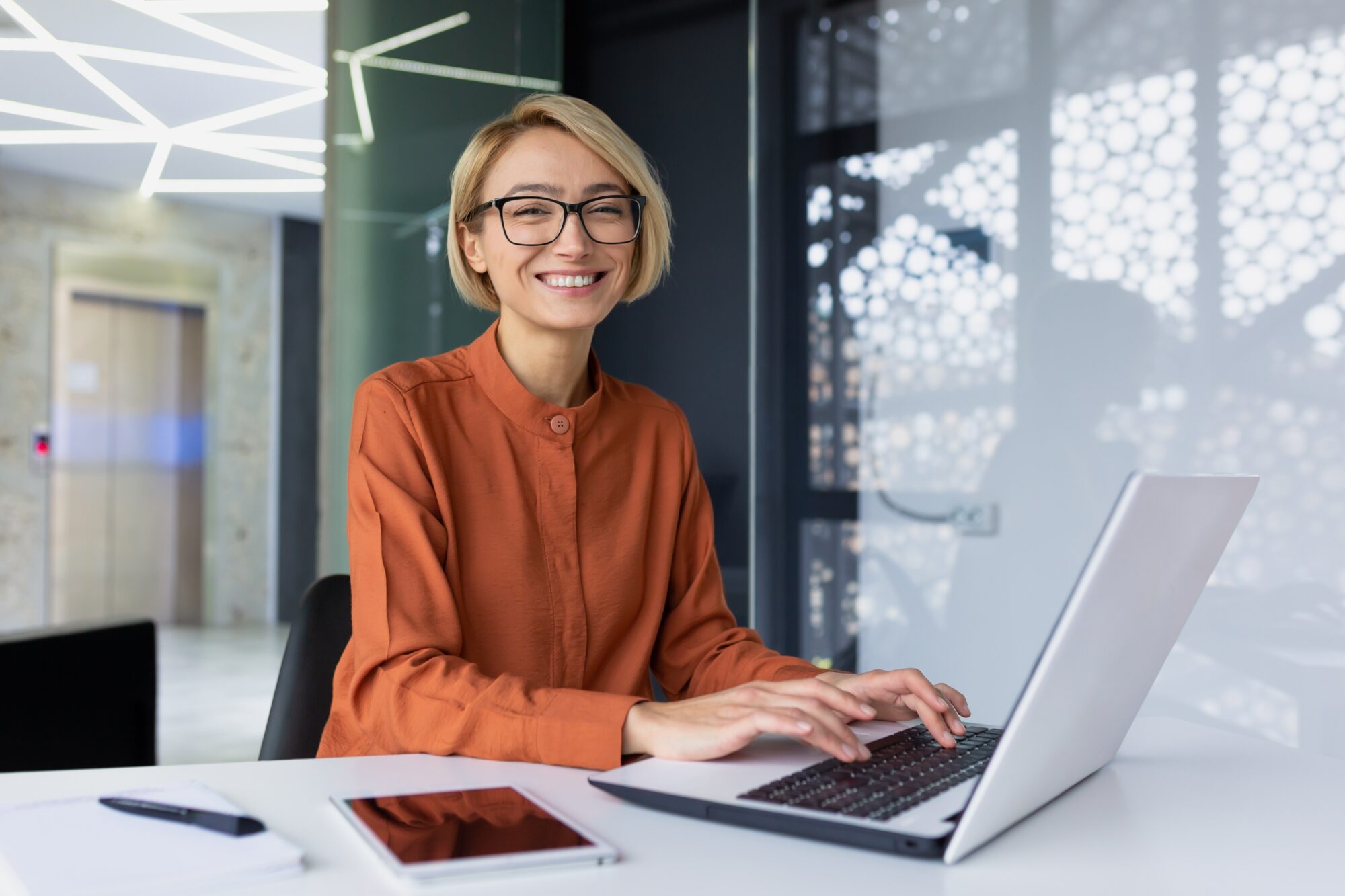 A woman with short blonde hair and glasses is sitting at a white desk, smiling at the camera. She is wearing an orange blouse and typing on a laptop focused on single pane of glass security. A notepad and tablet are on the desk, set against an office backdrop.