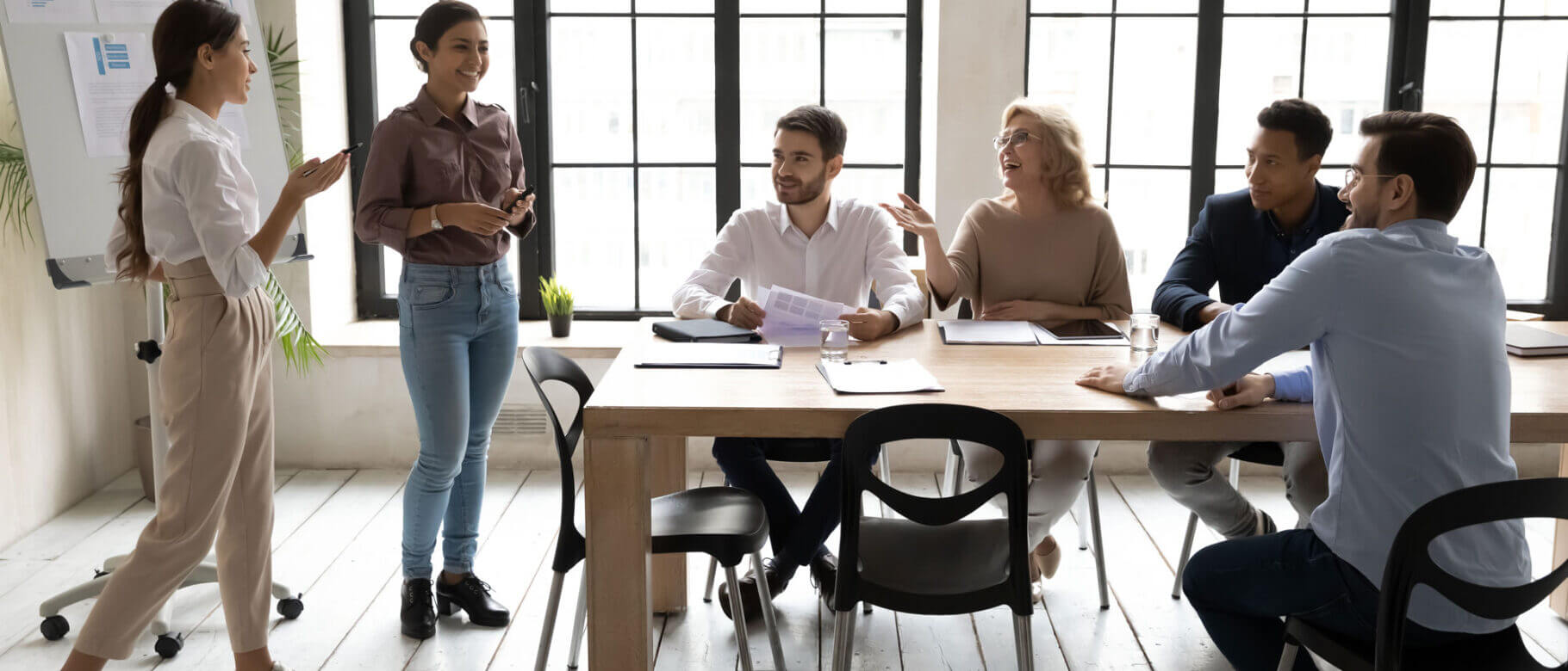 A group of six people are engaged in a lively discussion in a well-lit conference room. Two individuals stand near a whiteboard discussing non-proprietary access control hardware, while four others are seated at a table with papers and laptops. Large windows let in natural light.