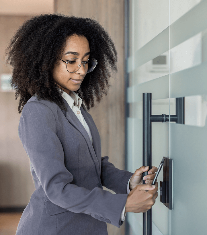Person with curly hair, wearing glasses and a gray suit, holds a smartphone or access card near an electronic lock on a glass door, appearing to unlock it. The setting looks like an office environment.