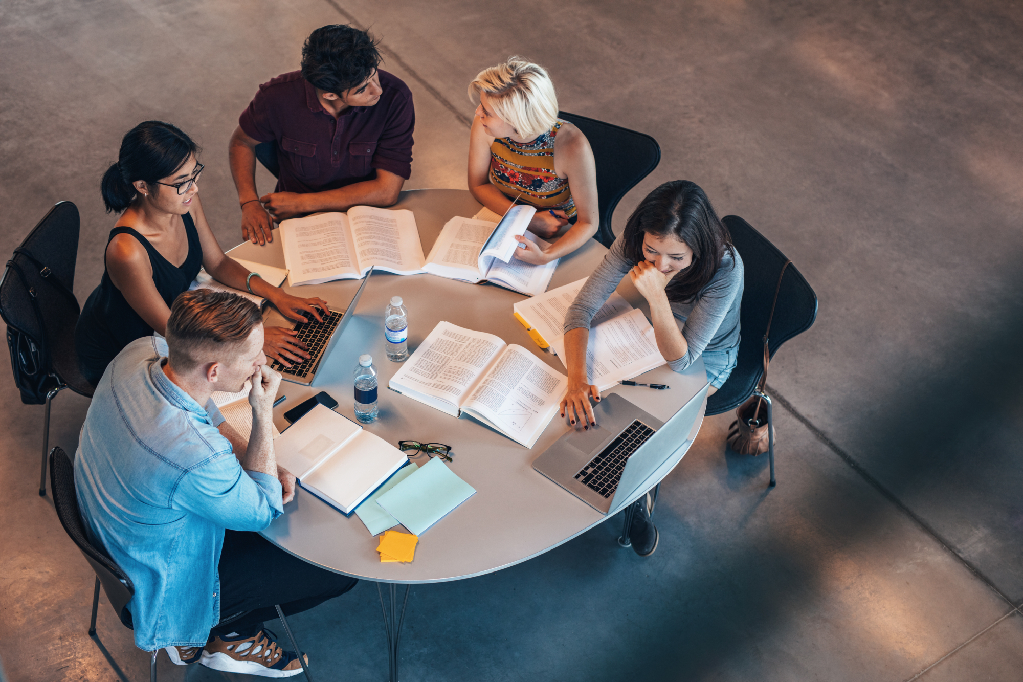 Five young adults sit around a round table with laptops, notebooks, and books, studying or discussing Shooter Detection technology in a modern, open space. Bottled water and sticky notes are also on the table.