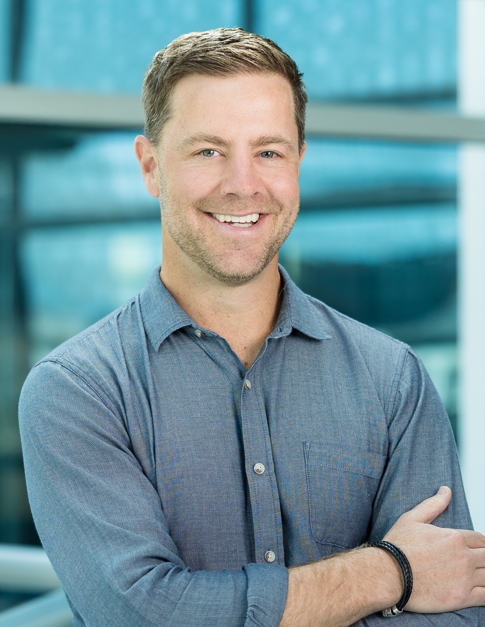 A smiling man with short brown hair and a beard, wearing a light blue button-up shirt, stands with arms crossed in front of a modern, glass-walled background—perfect for an about Genea introduction.