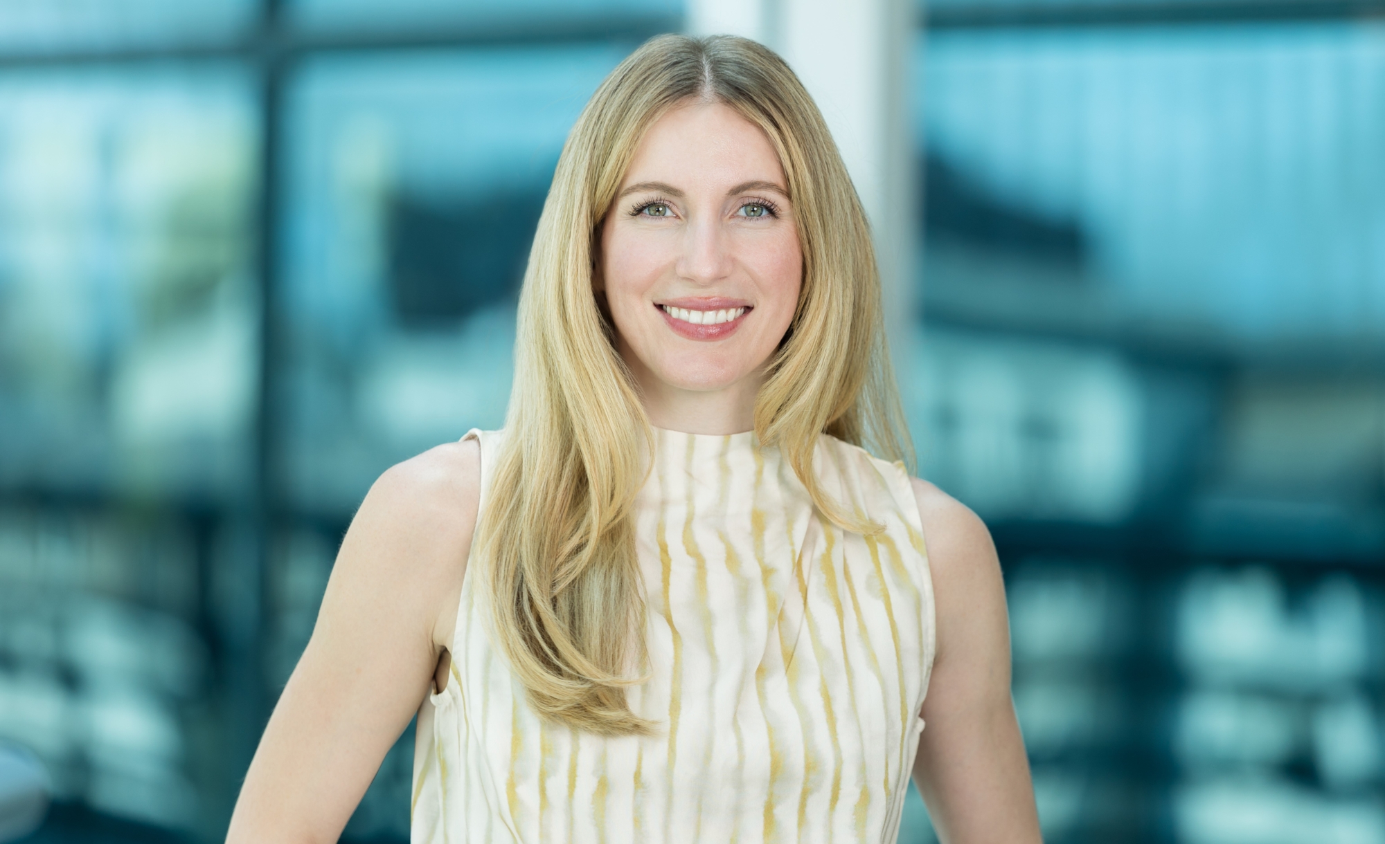 A woman with long blonde hair, wearing a sleeveless, light-colored top with a subtle striped pattern, smiles at the camera. The blurred blue and glass background suggests a modern indoor setting—perfect for an about Genea profile.