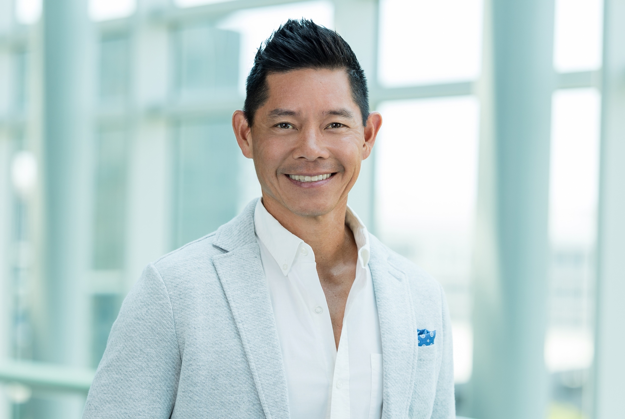 A man with short dark hair, wearing a light gray blazer over a white shirt, smiles while standing in a bright, modern building with large windows in the background, reflecting the innovative spirit about Genea.