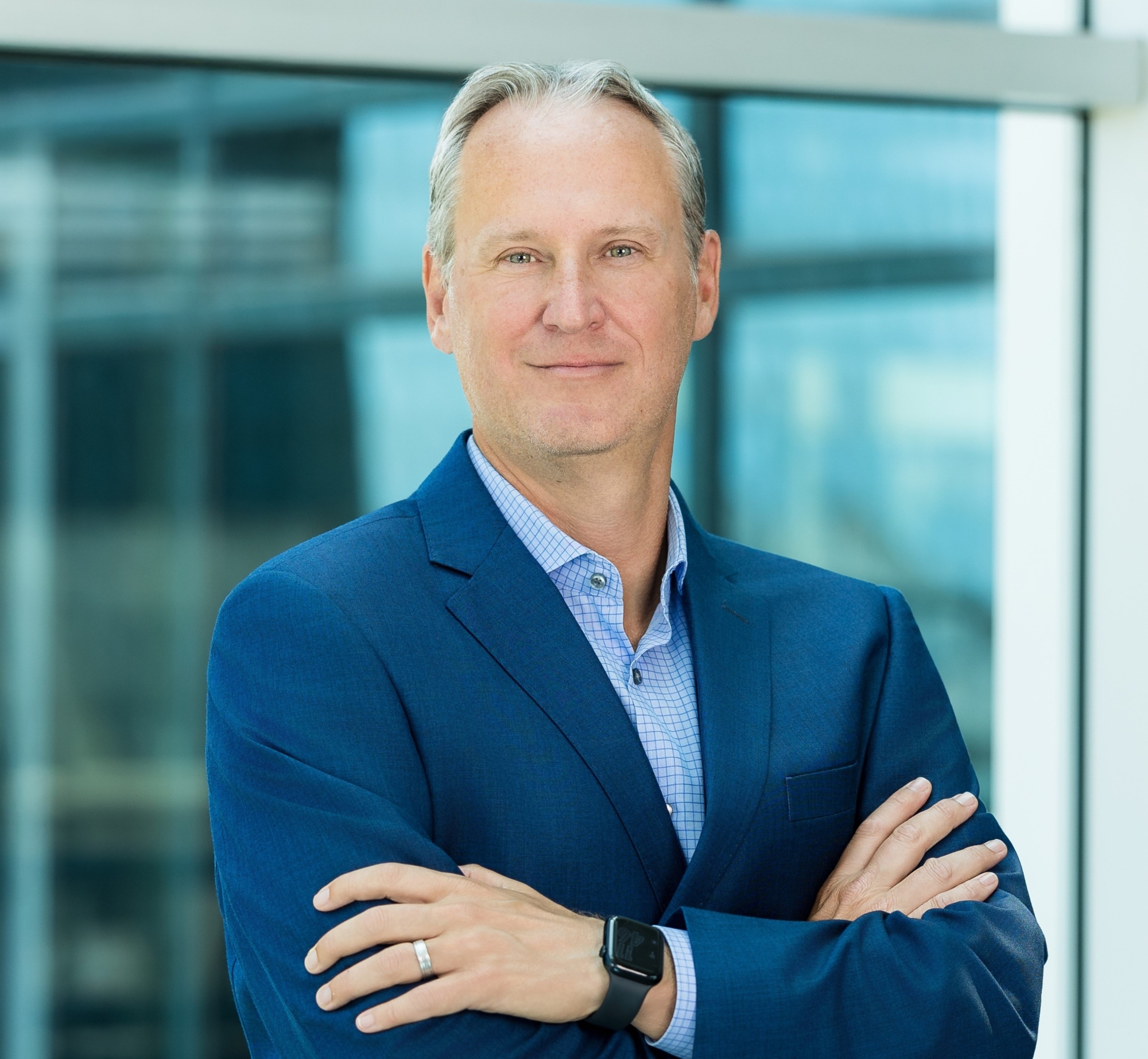 A middle-aged man with short gray hair wearing a blue suit, checked shirt, and smartwatch stands with arms crossed, smiling confidently in a modern office setting with large glass windows—reflecting his role at Genea.