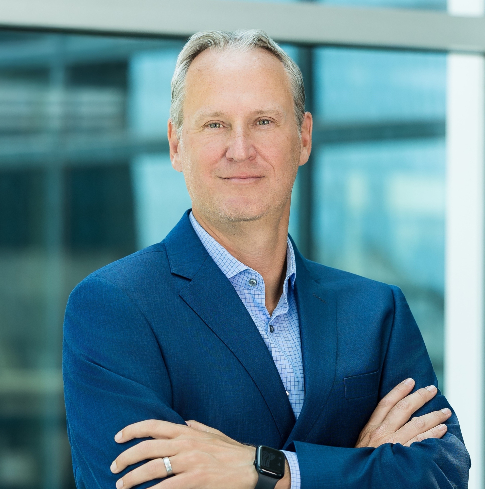 A middle-aged man with short gray hair wearing a blue suit and checked shirt stands with arms crossed, smiling slightly. He is in a modern office with large windows, reflecting the professional atmosphere at Genea.