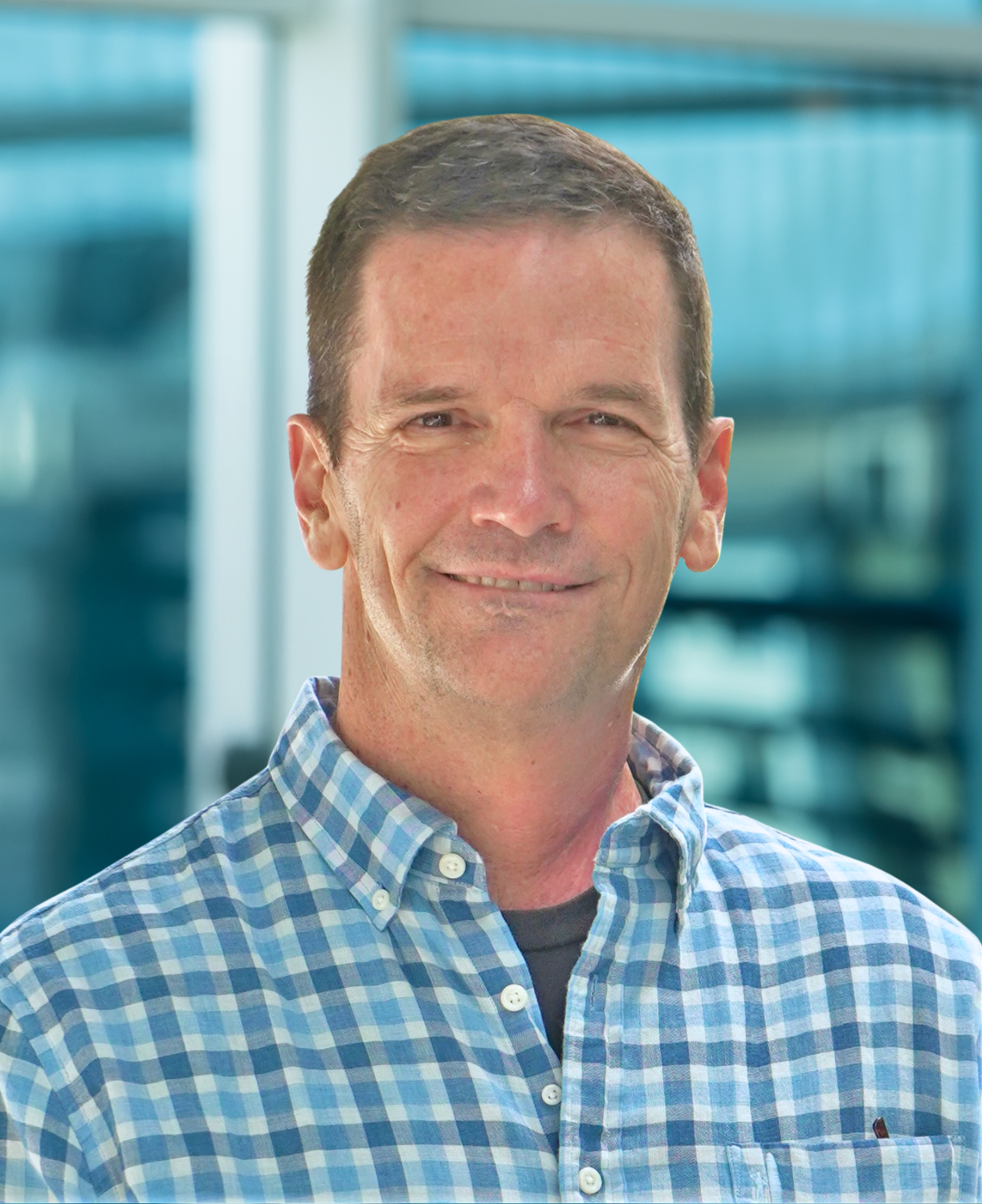 A man with short brown hair wearing a blue and white plaid shirt stands smiling in front of a blurred modern glass building background, reflecting the welcoming spirit found at about Genea.