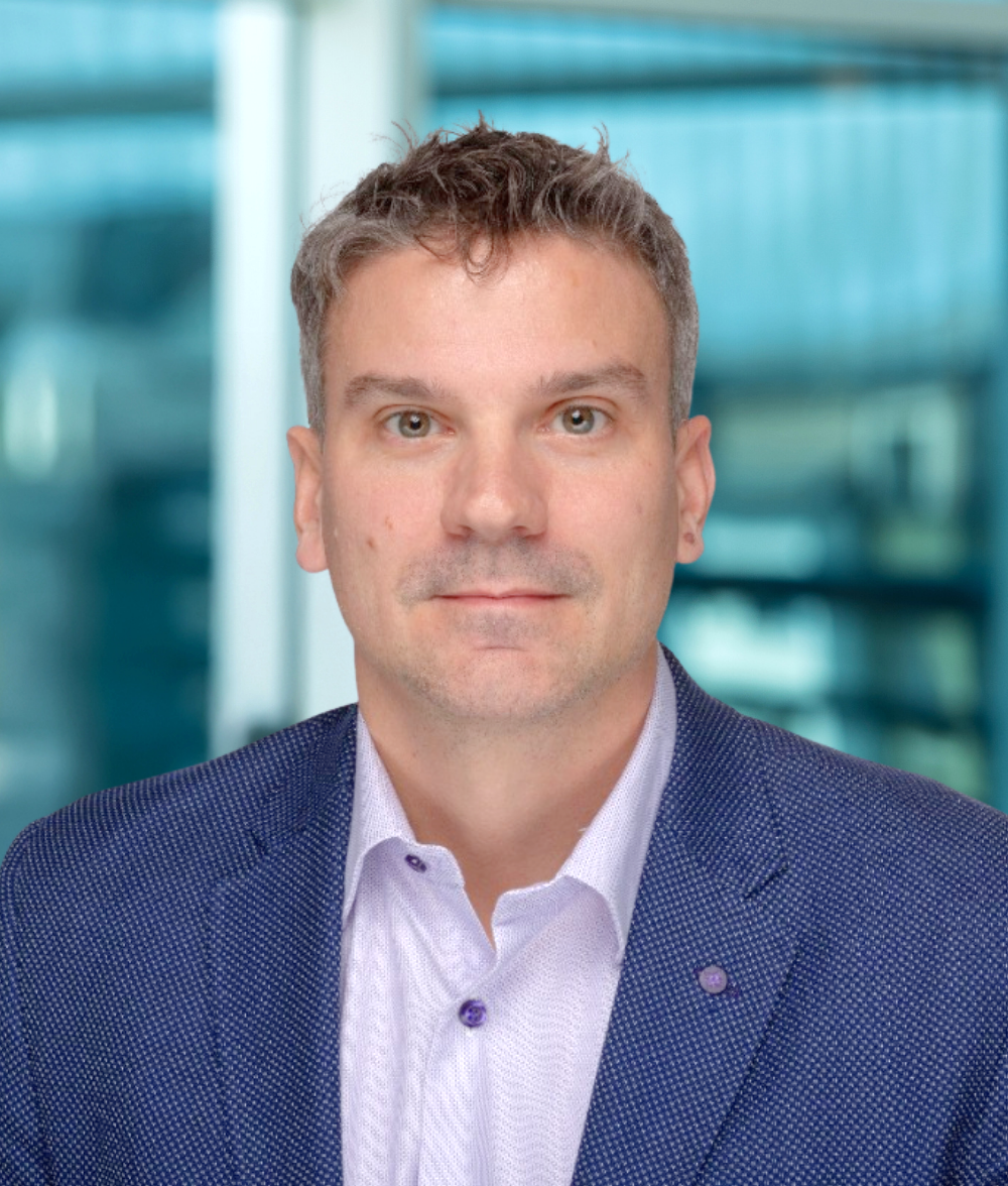 A man with short, curly gray hair, wearing a blue textured blazer over a light purple button-up shirt, sits in front of a blurred, modern glass background—reflecting a professional image about Genea.