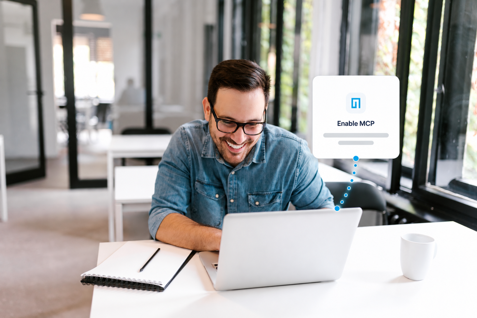 A man wearing glasses and a denim shirt smiles at his laptop in a bright office. A notepad and mug are on the table. A pop-up on his screen reads Enable MCP shooter detection.