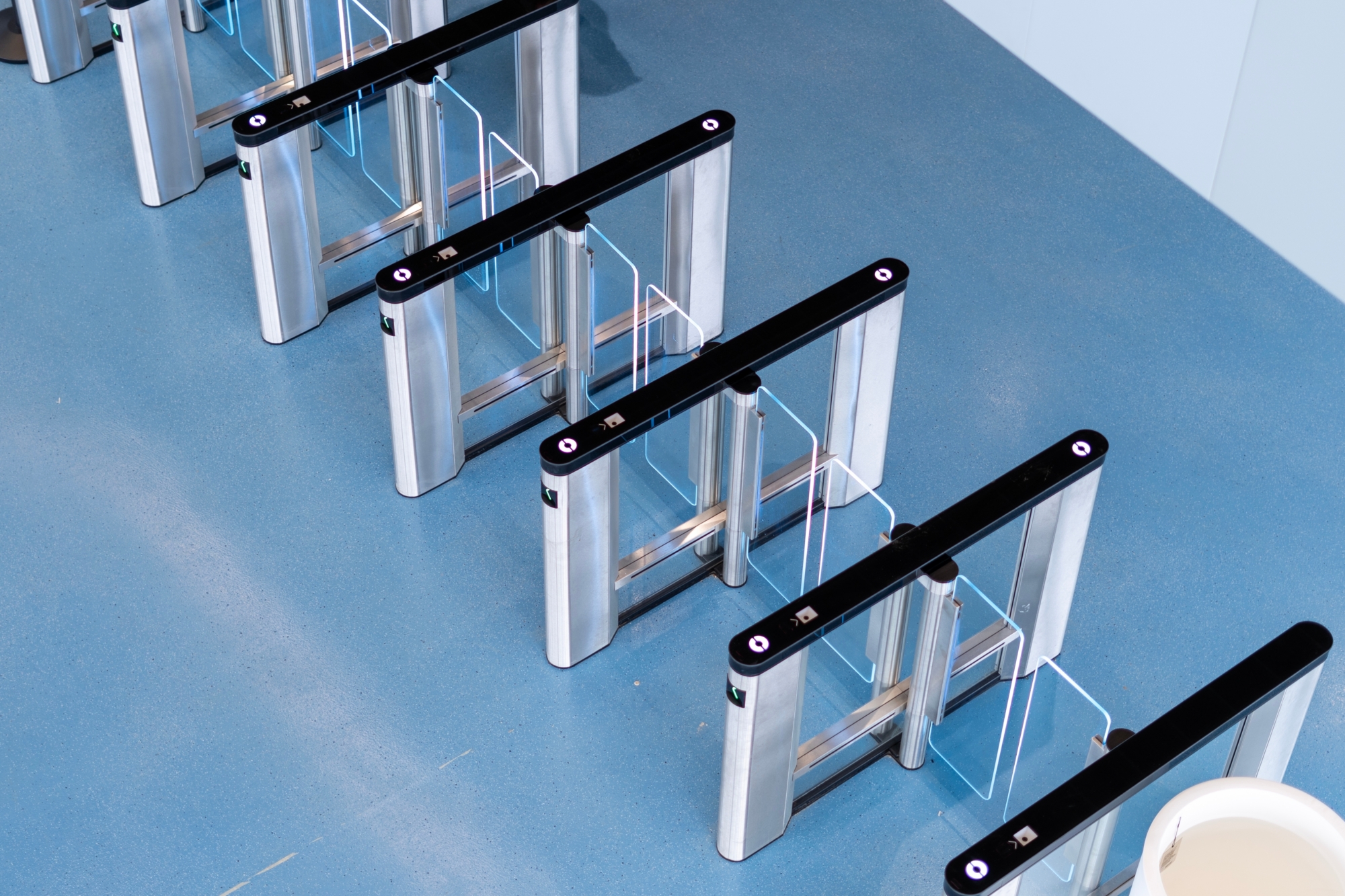 A row of modern security turnstiles with glass barriers on a blue floor, viewed from above. Designed for seamless access control migration, the turnstiles have black tops with circular lights and transparent glass panels.